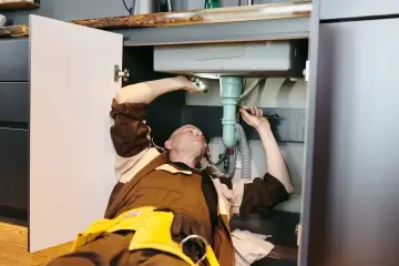 Hydraulik naprawia syfon pod zlewem, gdzie widać zakamienione rury ciepłej wody.