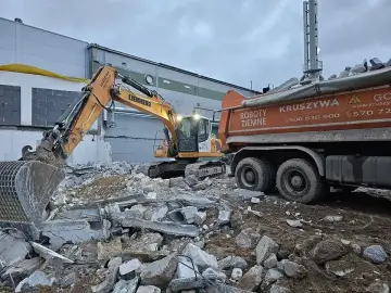 Żółty koparka pracuje przy rozbiórce gruzowiska. Obok stoi pomarańczowa wywrotka, gotowa zabrać urobek.