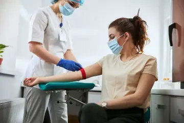 Pielęgniarka w maseczce i rękawiczkach zakłada opaskę uciskową na ramię pacjentki, przygotowując ją do pobrania krwi. Może to być związane z podwyższonym ob.