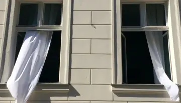 Weiße Gardinen wehen aus zwei offenen Fenstern eines hellen Gebäudes. Die Frage "Gardinen waschen wieviel Grad?" beschäftigt die Hausfrau.