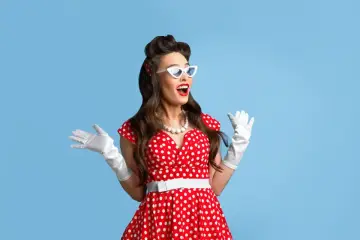 Urocza pin up girl w czerwonej sukience w grochy, białych rękawiczkach i okularach w kształcie kocich oczu, z uniesionymi rękami w geście zdziwienia.