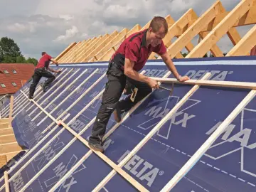 Dwaj budowlańcy pracują na dachu, układając drewniane łaty na folii wstępnego krycia.