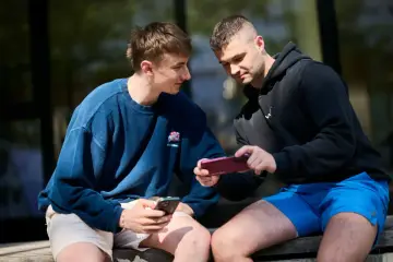 Dwóch młodych mężczyzn siedzi na ławce, jeden pokazuje drugiemu, jak przesłać zdjęcia z telefonu na telefon.