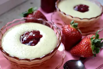 Dwa pucharki z kremowym budyniem jaglanym, ozdobione konfiturą z owoców leśnych, obok świeże truskawki i łyżeczka.