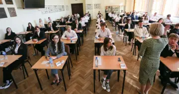 Uczniowie siedzą w ławkach podczas egzaminu, a nauczycielka stoi z tyłu.
