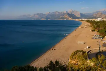 Słoneczna plaża na riwierze tureckiej z turkusowym morzem i górami w tle. Kilka osób pływa, inni odpoczywają na piasku.