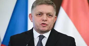 Premier Słowacji w garniturze i krawacie, przemawia na tle flag.
