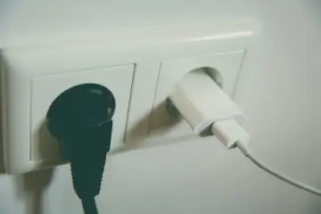 Dwa gniazdka elektryczne: jedno z czarną wtyczką, drugie z białą ładowarką. Czekają na napięcie w gniazdku.