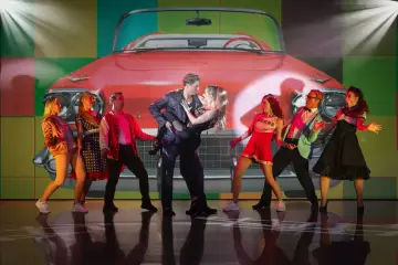 Tänzer in 50er-Jahre-Outfits vor einem roten Oldtimer-Cabriolet. Ein Paar tanzt im Zentrum, umgeben von anderen Darstellern. Ein lebendiger Moment aus Musicals Deutschland.