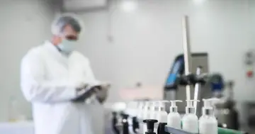 Pracownik w białej odzieży ochronnej nadzoruje linię produkcyjną z buteleczkami zawierającymi kosmetyki, gdzie kluczowym składnikiem jest fenoksyetanol.
