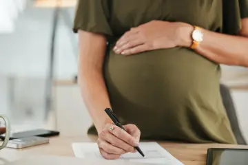 Kobieta w ciąży wypełnia dokumenty, planując, kto płaci za macierzyński. Na biurku telefon, kalkulator i tablet.