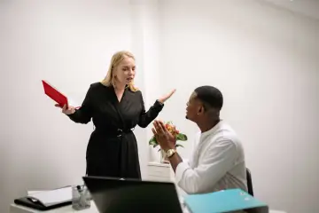Une femme en colère, tenant un livre rouge, semble se faire insulter au travail par un collègue qui a les mains jointes.