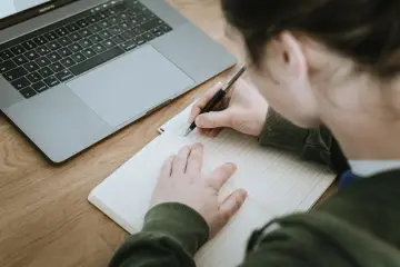 Uczeń pisze w zeszycie, obok leży laptop. Nauczyciel przygotowuje opinię o uczniu.