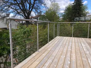 Nowe, drewniane deski tarasowe i solidne mocowanie balustrady balkonowej ze stali nierdzewnej. W tle zieleń drzew i błękitne niebo.
