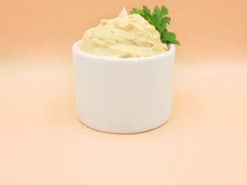 Puszysty hummus z ciecierzycy, udekorowany gałązką natki pietruszki, w białej miseczce.
