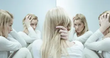 Kobieta z blond włosami w białej bluzie, z rękami na głowie, otoczona odbiciami w lustrze, symbolizującymi walkę z nerwicą lękową, być może szukająca ulgi w kwetaplex.