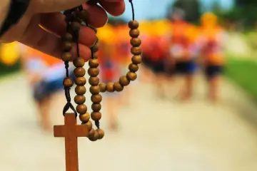 Drewniany różaniec z krzyżem w dłoni, symbolizujący modlitwę za brata. W tle rozmyte sylwetki ludzi.