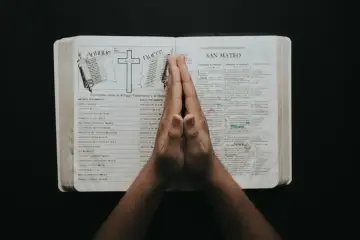 Dłonie złożone do modlitwy nad otwartą Biblią, na stronach której widnieje porównanie Starego i Nowego Testamentu, a także fragment Ewangelii wg św. Mateusza. W tle widać symbol krzyża i zwoju, nawiązujące do treści. To chwila refleksji, być może nad p...