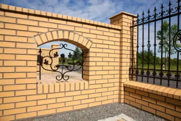Eleganckie ogrodzenia z klinkieru wzory: żółta cegła, łukowe przejście z kutą ozdobą i czarna, metalowa brama.