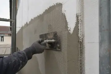 Ręka w rękawicy zatapia siatkę w tynku, tworząc wzmocnienie elewacji.