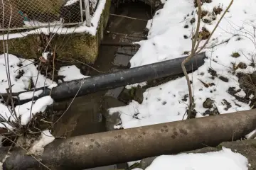 Dwie rury biegną nad strumieniem. Woda w rurach zamarznięta, otoczenie pokryte śniegiem.
