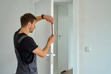 Mężczyzna w ogrodniczkach sprawdza poziomem, jak wstawić drzwi. Precyzyjne pomiary to klucz do sukcesu.