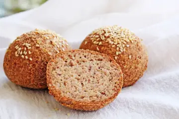 Trzy keto bułki posypane sezamem, jedna przekrojona na pół, na białym tle.