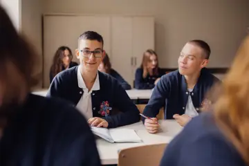Uczniowie liceum podczas lekcji. Chłopcy w mundurkach siedzą przy ławkach, jeden z nich uśmiecha się do aparatu, drugi pisze.