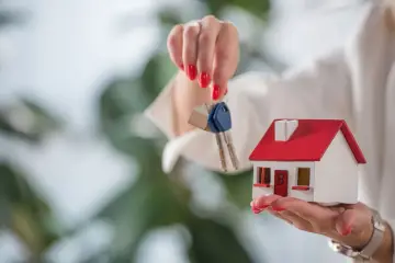 Kobieta trzyma w dłoniach miniaturowy domek i klucze. To symboliczne przedstawienie, jak mieszkanie może stanowić wkład własny przy zakupie nieruchomości.