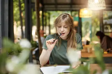 Młoda kobieta zamyślona nad notatkami, gryzie długopis. Może pisze esej "for and against", szukając argumentów.