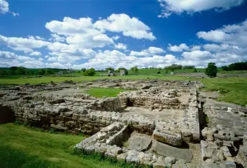 Extensive roman ruin spread across a green field under a blue sky with fluffy clouds.