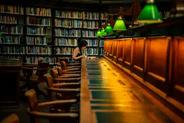 Dziewczyna uczy się w bibliotece, otoczona regałami pełnymi książek. To miejsce, gdzie często spędzają czas uczniowie, przygotowując się do lekcji i sprawdzianów.