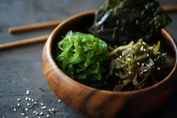 Trzy rodzaje jadalnych wodorostów: zielone wakame, ciemne kombu i sałatka z wodorostów z sezamem, w drewnianej misce.