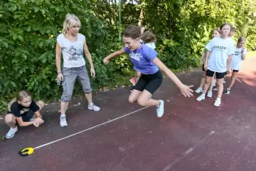 Dziewczynka skacze przez linię podczas testów sprawnościowych w klasach 1-3. Nauczycielka obserwuje.