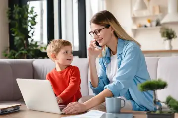 Mama na urlopie wypoczynkowym po wychowawczym pracuje przy laptopie, a synek patrzy na nią z zaciekawieniem.