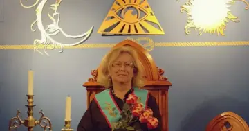Kobieta w loży masońskiej, siedząca na ozdobnym tronie, z różami w dłoni. W tle symbole: oko opatrzności, słońce i księżyc.