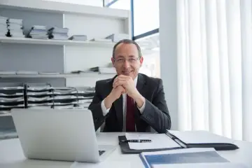 Uśmiechnięty przedsiębiorca w garniturze siedzi przy biurku z laptopem i dokumentami.