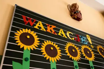 Kolorowe litery "WAKACJE!" i słoneczniki na tablicy zapowiadają radosne zakończenie roku szkolnego.
