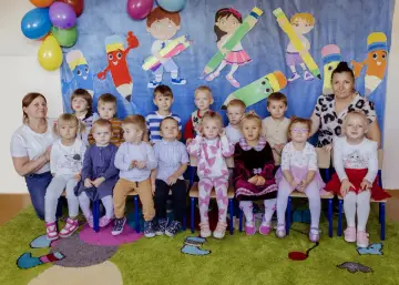 Grupa przedszkolaków z panią, w tym Tymon i Tymoteusz, pozuje do zdjęcia na tle kolorowych kredek.