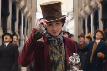 Timothée Chalamet w filmach, jako młody Willy Wonka, w cylindrze i barwnym stroju, z uśmiechem pozdrawia tłum.