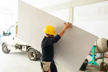 Budowlaniec w żółtym kasku niesie dużą płytę gk. Obok stoi betoniarka i samochód dostawczy.