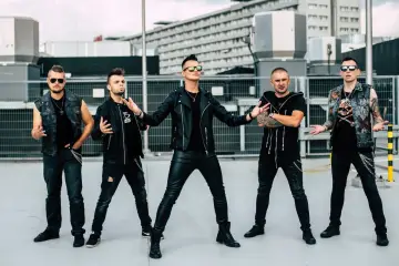 Pięciu mężczyzn w czarnych, rockowych strojach, jakby nocny kochanek o jeden most za daleko, pozuje na tle miejskiej architektury.