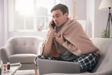 Chory mężczyzna owinięty kocem rozmawia przez telefon, szukając informacji, jak obliczyć świadczenie chorobowe.