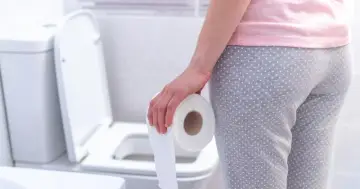 Osoba w piżamie trzymająca rolkę papieru toaletowego przy toalecie. Może szukać ulgi, np. po zastosowaniu laxigen.