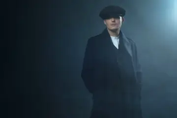 Mężczyzna w kaszkiecie i płaszczu, z charakterystycznym wąsem, emanuje chłodnym spojrzeniem w stylu Peaky Blinders.