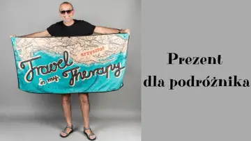 Uśmiechnięty mężczyzna w okularach prezentuje ręcznik z mapą świata i napisem "Travel is my therapy". Idealny prezent dla podróżnika.