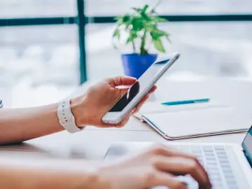 Dłoń trzyma smartfon, korzystając z tetheringu, podczas gdy druga ręka pisze na laptopie. Obok leży notatnik i długopis.