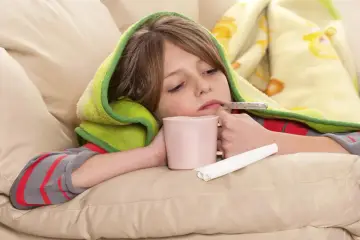 Dziecko z gorączką, pod kocykiem, z termometrem w ustach i kubkiem w dłoni. To typowe objawy grypy u dzieci.