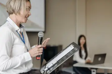 Kobieta z mikrofonem, emanująca charyzmą, przemawia do publiczności. W tle siedzi słuchaczka z laptopem.