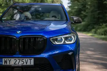 Błyszczący, niebieski BMW X5 M Competition na drodze leśnej. Charakterystyczne reflektory i czarny grill podkreślają jego sportowy charakter.